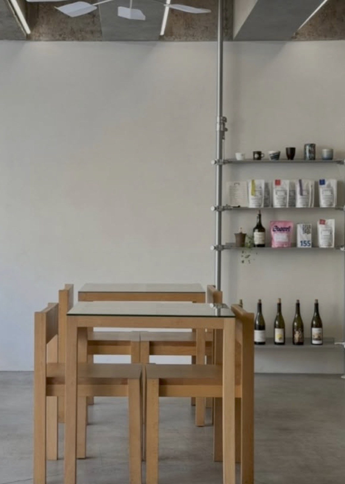A wooden dining table with glass top beside a shelf of natural wine bottles, ceramics and bagged coffee