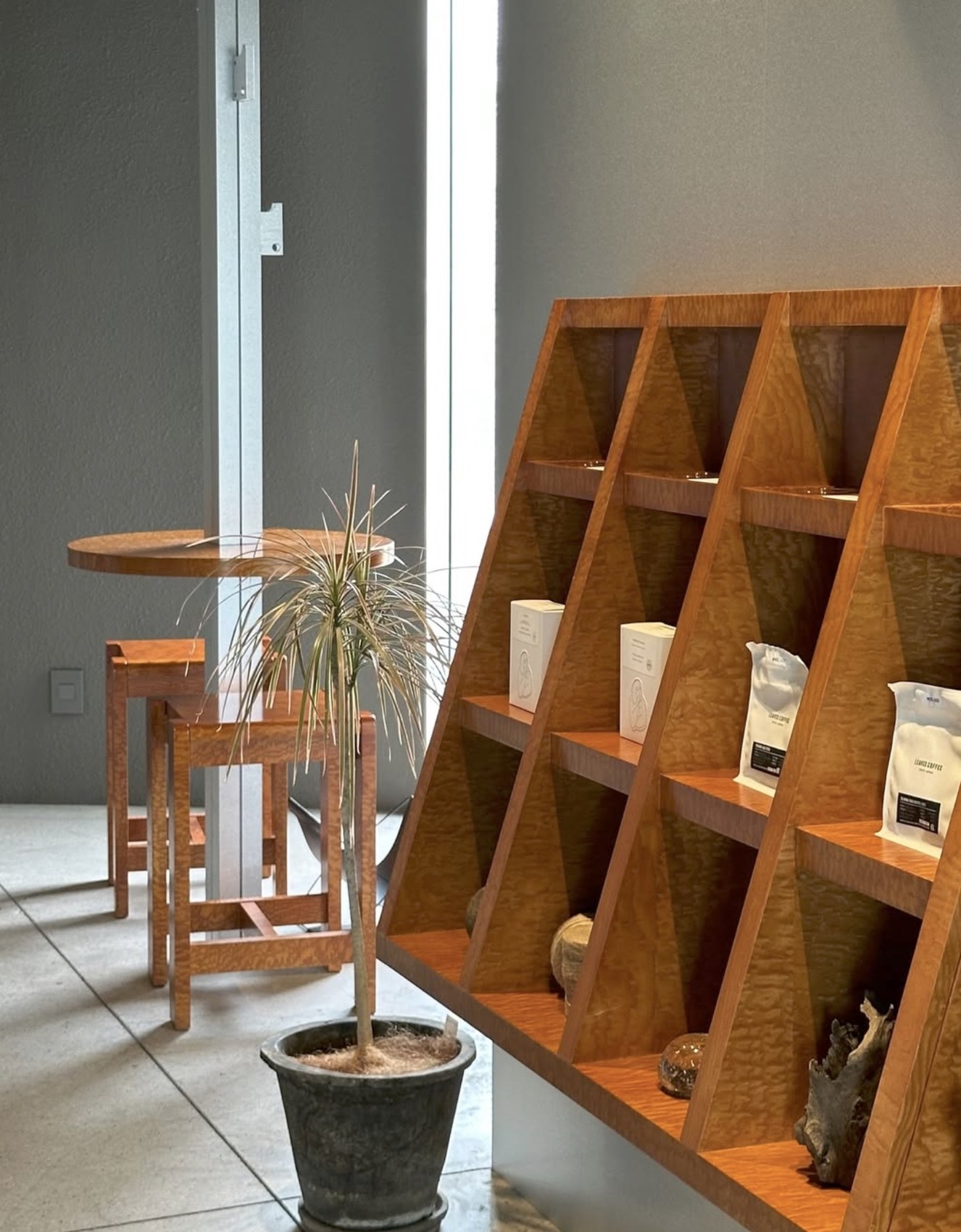 A triangular wooden shelving unit displaying retail coffee bags and boxes, a small round table and stools by the window behind