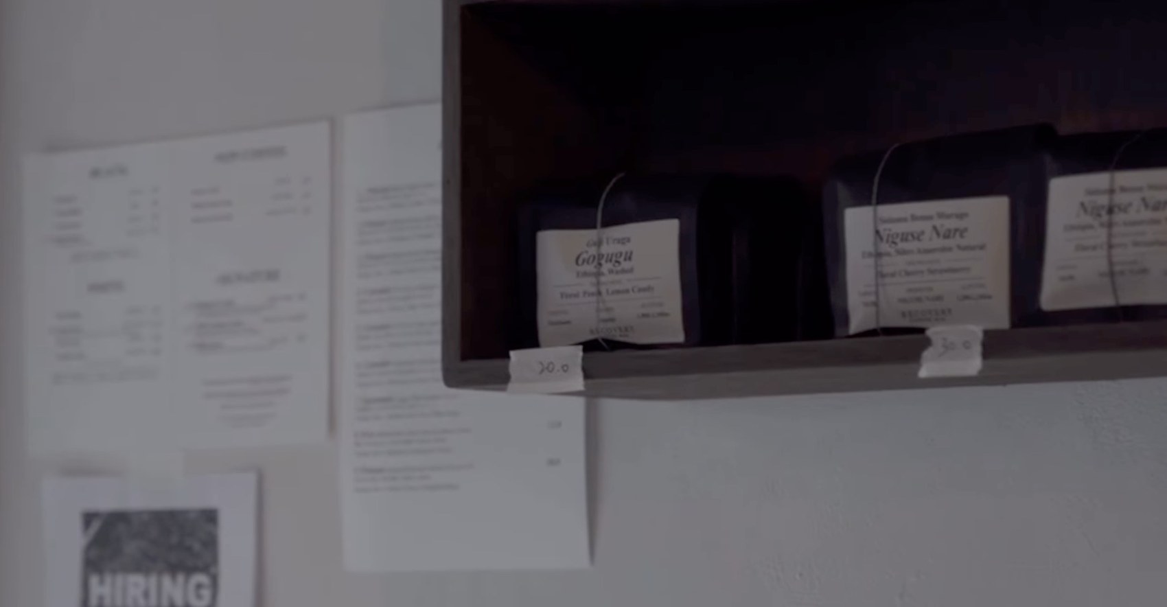 Bagged single origin beans labeled Gogugu and Niguse Nare on a dark wooden shelf above menus pinned to the wall