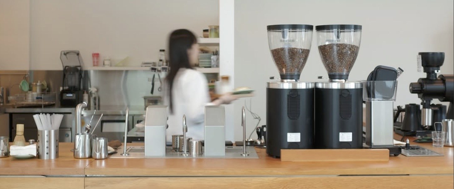 A pair of bean grinders on a wooden counter, a barista in motion behind them at the bar