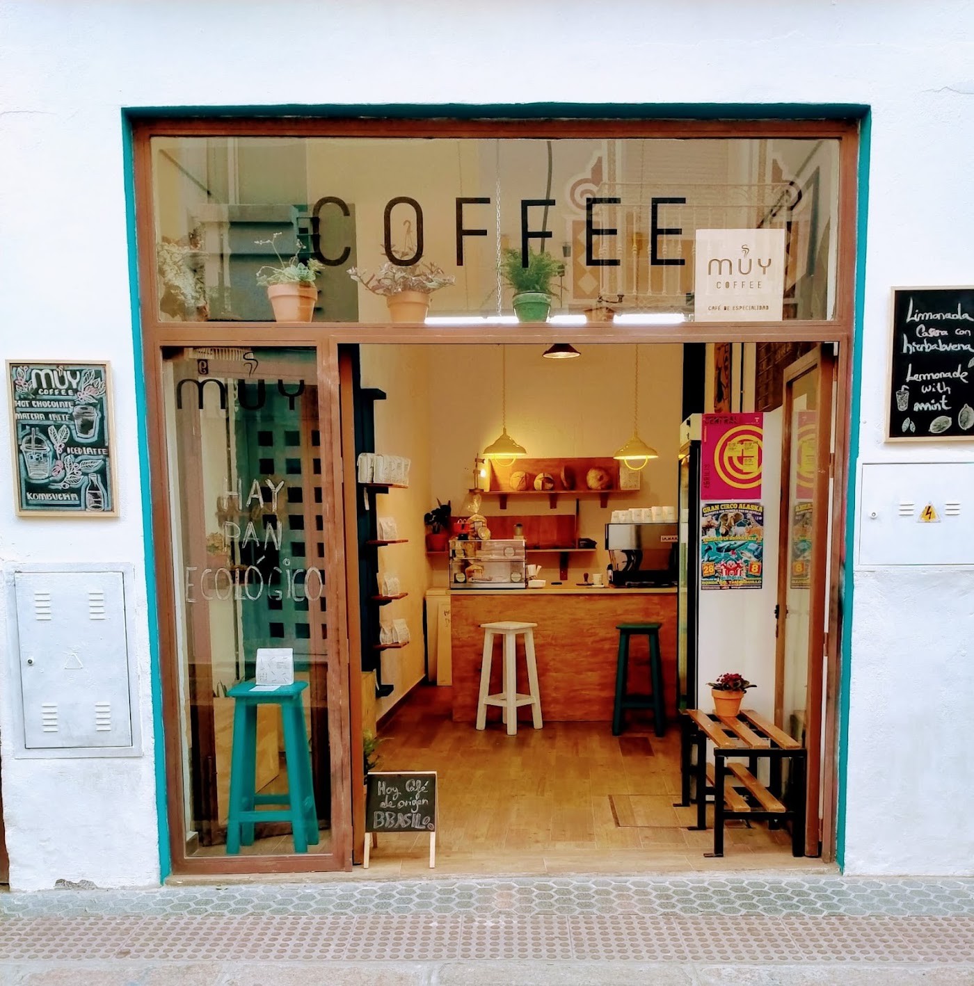 The empty MUY Coffee storefront with COFFEE lettering on the glass, teal stools, hanging bread shelves, and warm pendant lights
