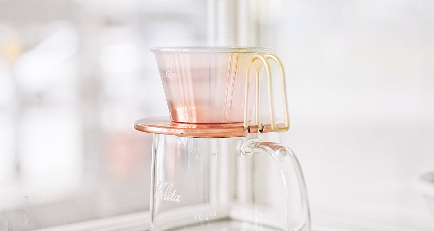 A pink ceramic Kalita Wave dripper with a gold handle set on a glass carafe at Apartment Coffee
