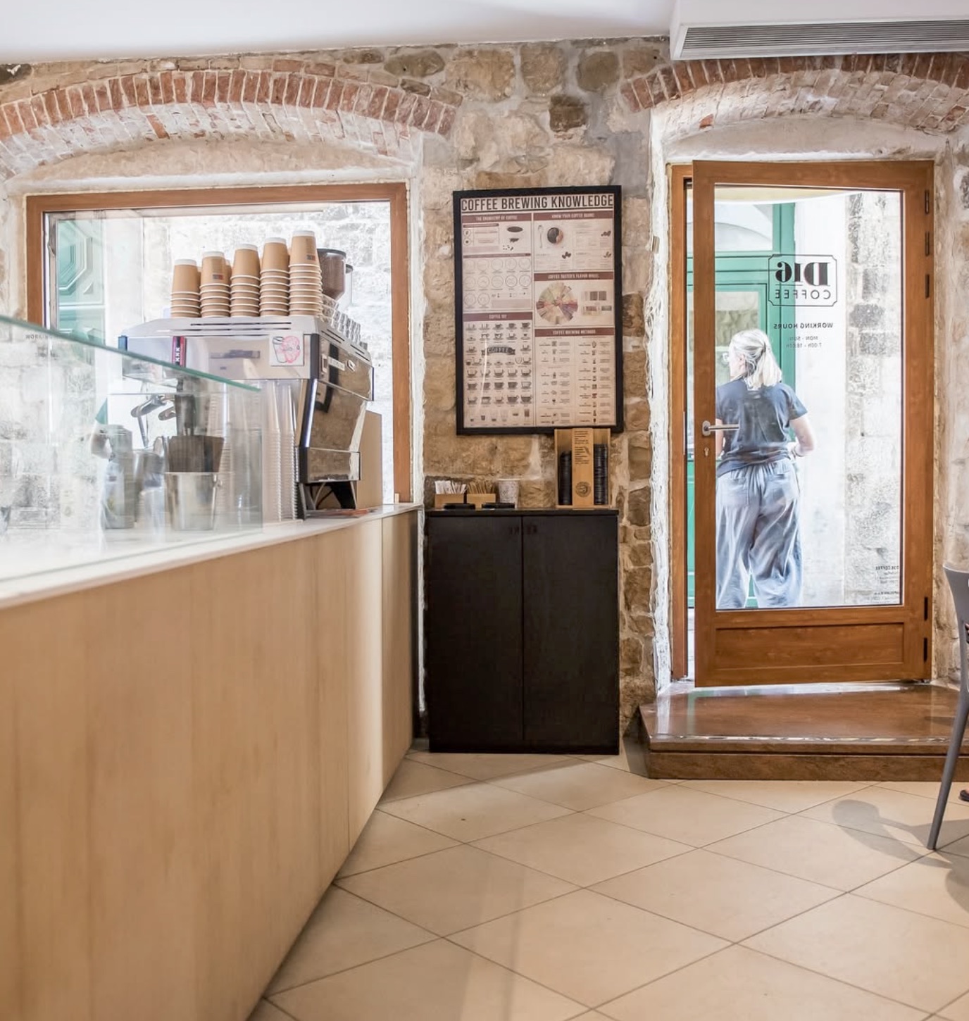 The stone-walled interior of D16 Coffee with an espresso machine and stacked takeaway cups on a pale wood counter, a coffee brewing chart on the wall, and a glass door bearing the cafe's logo