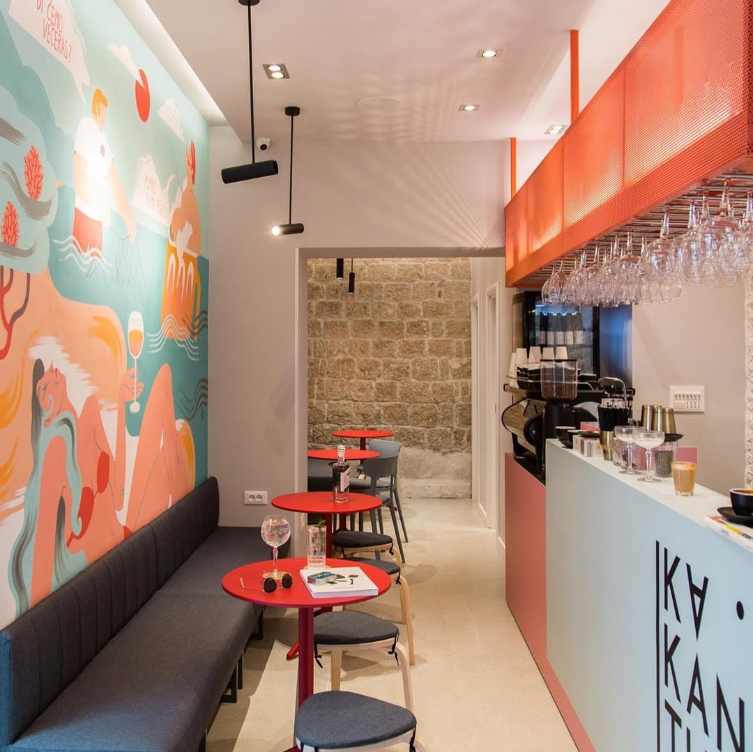 A narrow interior with a coastal mural, red bistro tables, and a glass topped bar at KaKantun