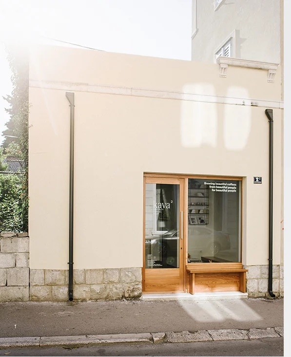 The sunlit cream facade of kava2 with a timber-framed glass door lettered 'kava2' and the line 'Brewing beautiful coffees from beautiful people for beautiful people', above a stone-block base on a quiet Split street