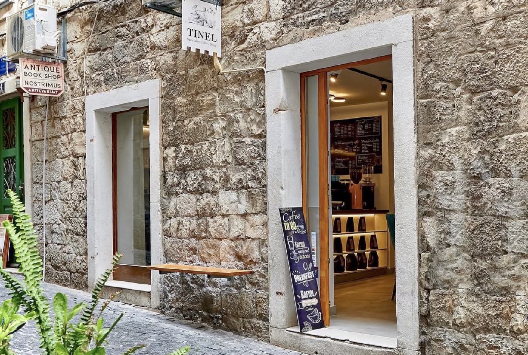 The stone facade of Tinel Specialty Coffee Shop in a Split alley, with a hanging TINEL sign, an open timber door showing the menu board and espresso setup inside, and a Coffee To Go chalkboard at the entrance