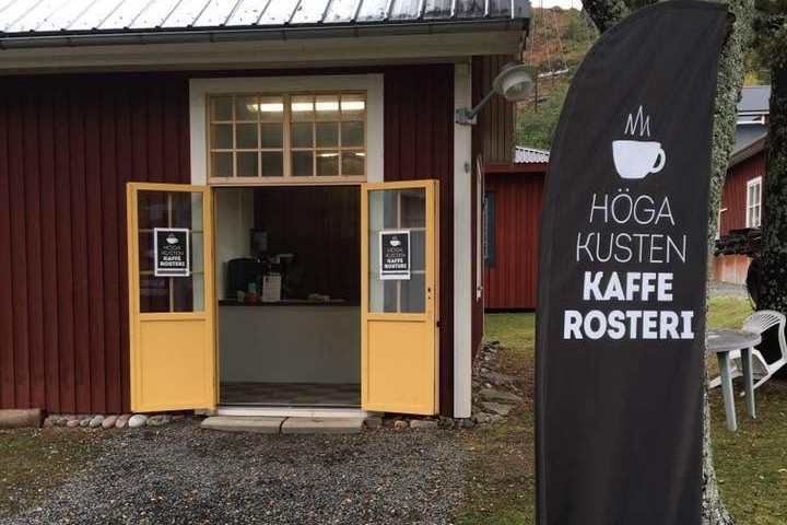 The red barn building with yellow doors and a HOGA KUSTEN KAFFE ROSTERI banner flag out front, coffee cup logo and phone number printed on the sign