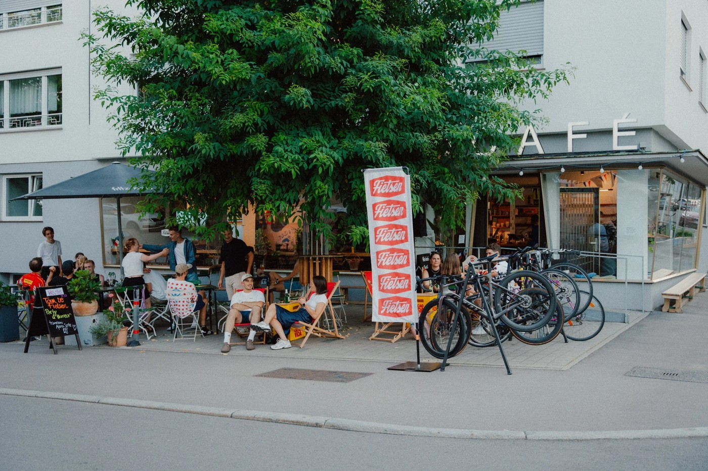 The corner storefront of Fietsen Radcafé under a large tree, with outdoor seating, a row of parked bicycles, and a banner reading Fietsen out front