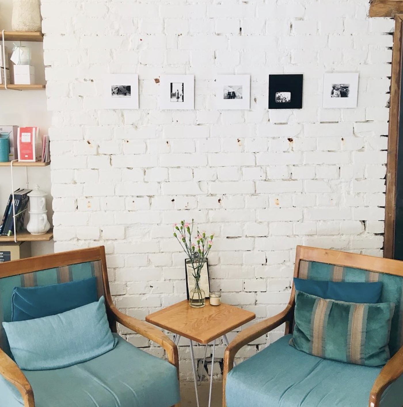 Two teal armchairs facing each other over a small round table with a vase of flowers, set against a white brick wall hung with framed black and white photos