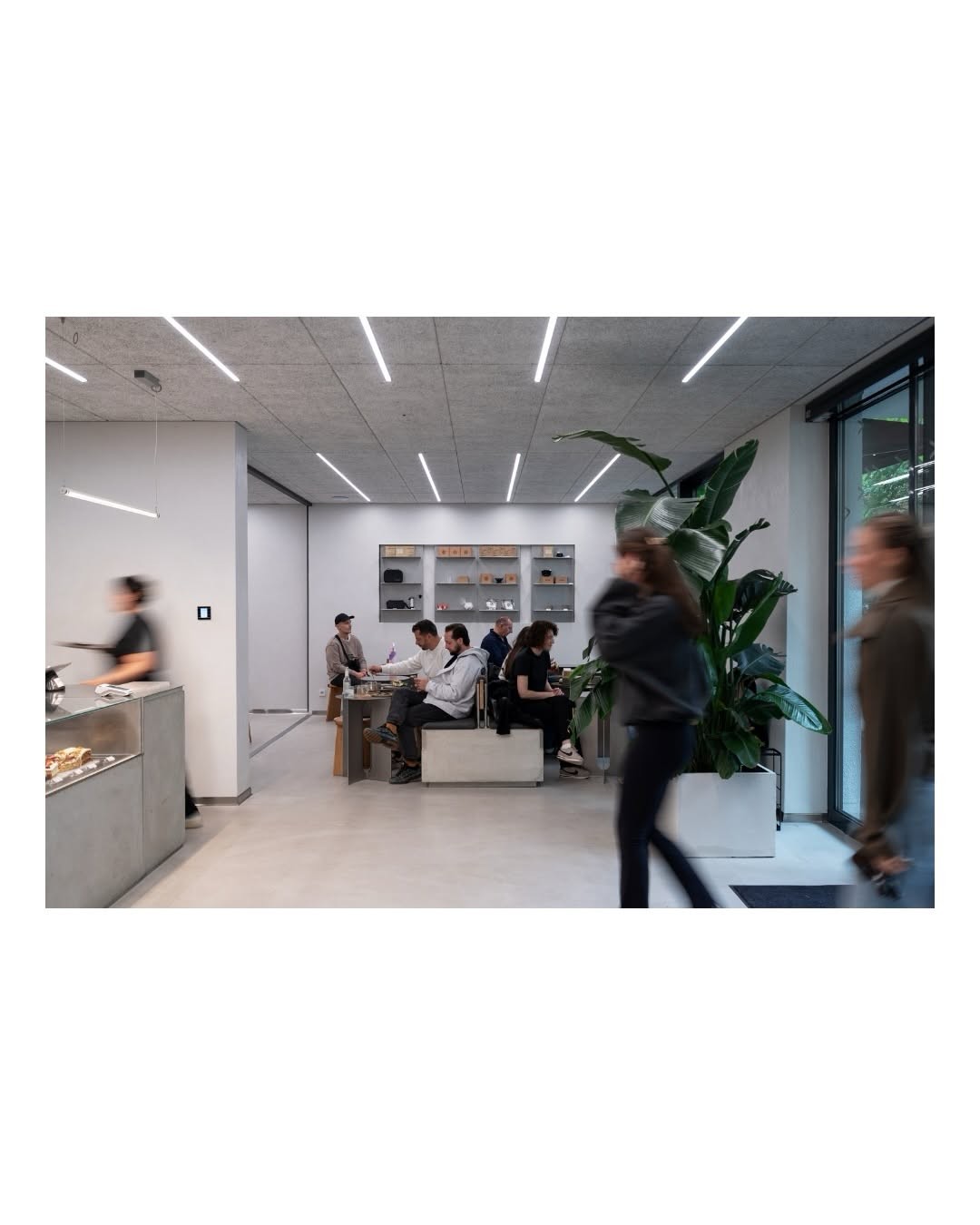 The interior of Taraba with a retail shelf, pastry case, and customers seated at a communal table, two people walking through the foreground in motion blur