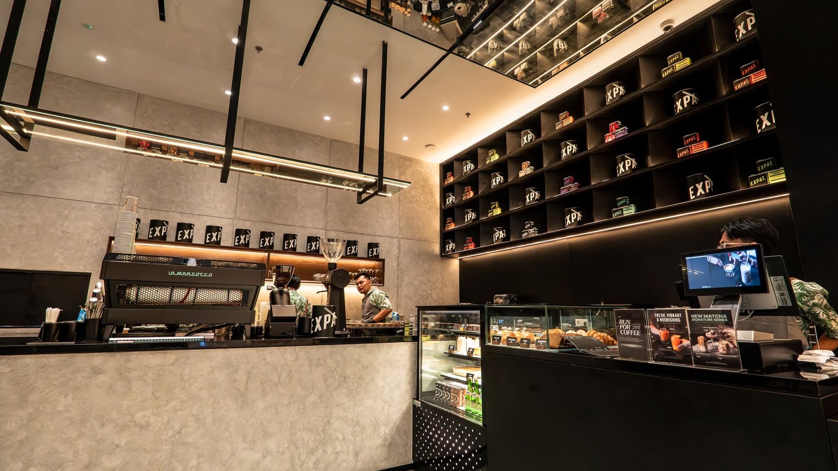 The Expat. Roasters counter with a La Marzocco espresso machine, shelves of branded retail cups and the pastry display case