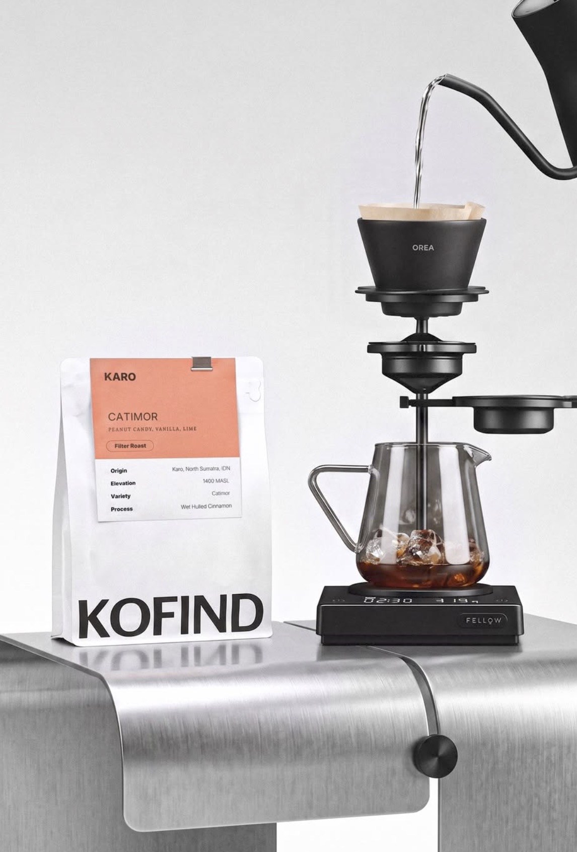 A bag of Kofind's Karo Catimor filter roast beans beside an Orea pour over dripper brewing iced coffee into a glass carafe