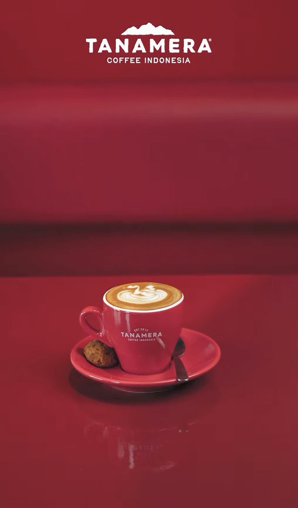 A latte with swan shaped latte art in a red branded Tanamera Coffee Indonesia cup and saucer with a small cookie