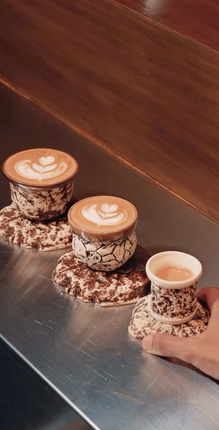 Three lattes with leaf shaped latte art in speckled ceramic cups on matching stone like coasters on a steel counter