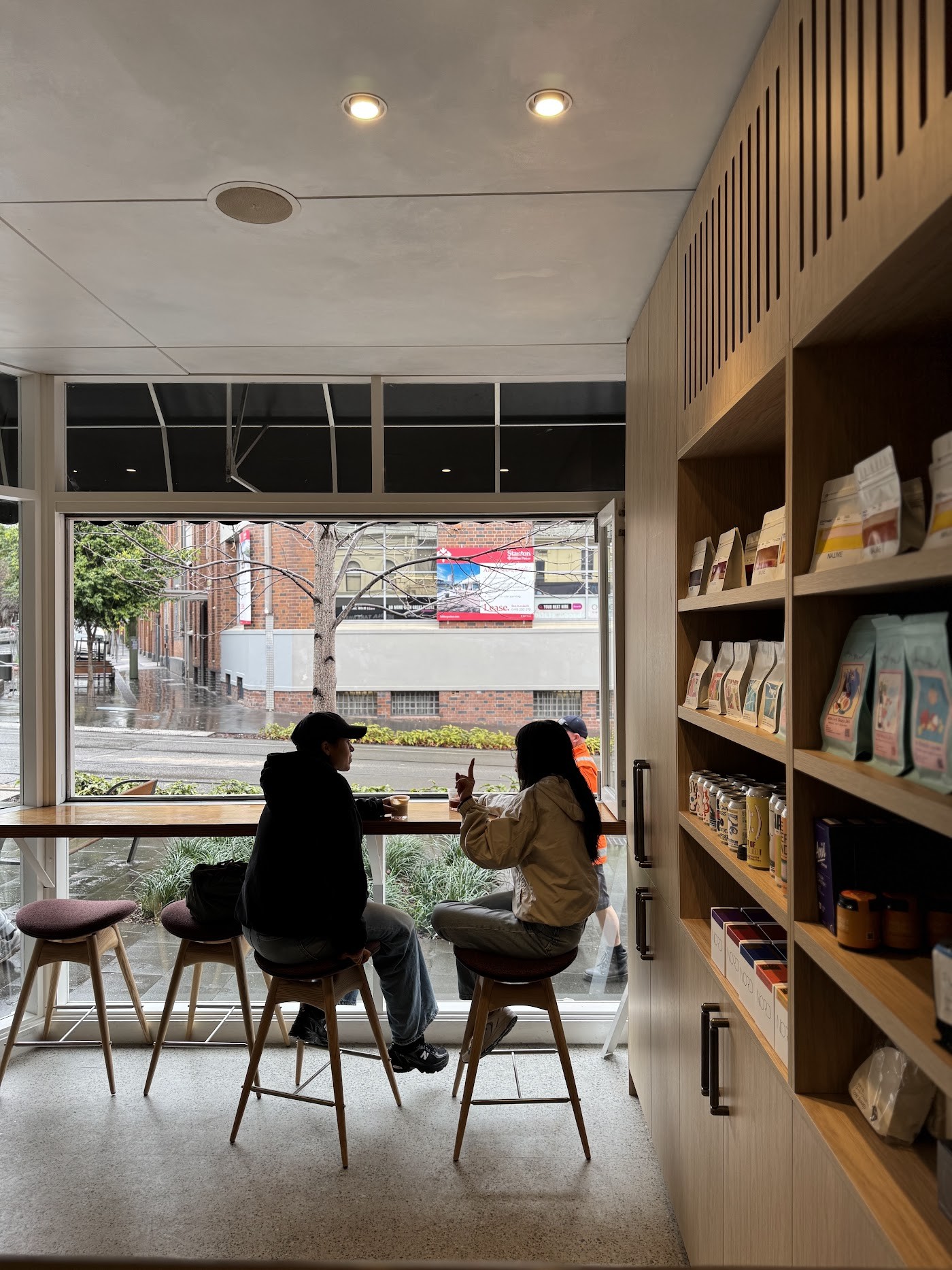 Two customers seated with their backs to the camera at Beta's window bar, beside a wall of retail coffee bags and cans
