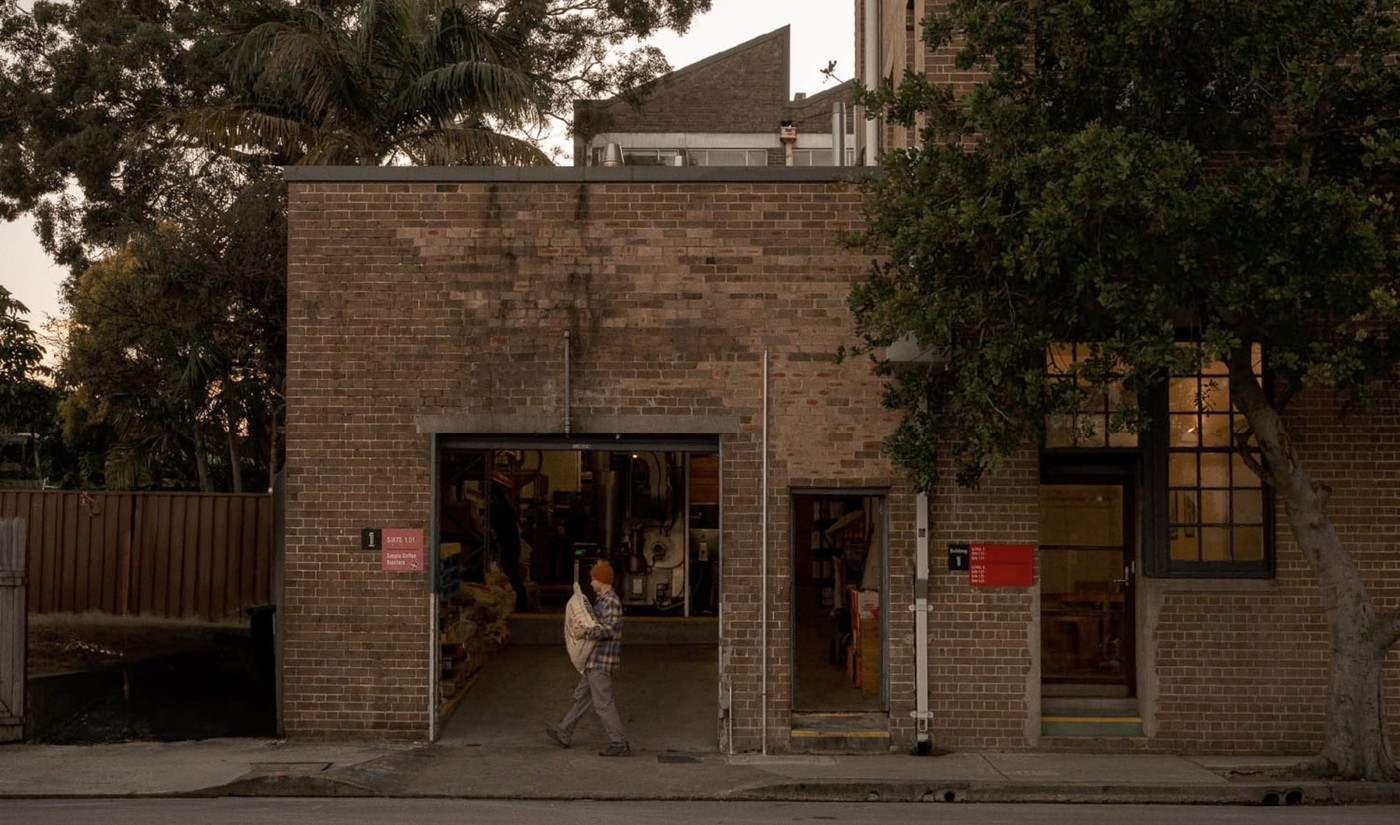 The brick exterior of Sample Coffee Roasters at dusk, a worker carrying a green coffee sack through the open roastery door
