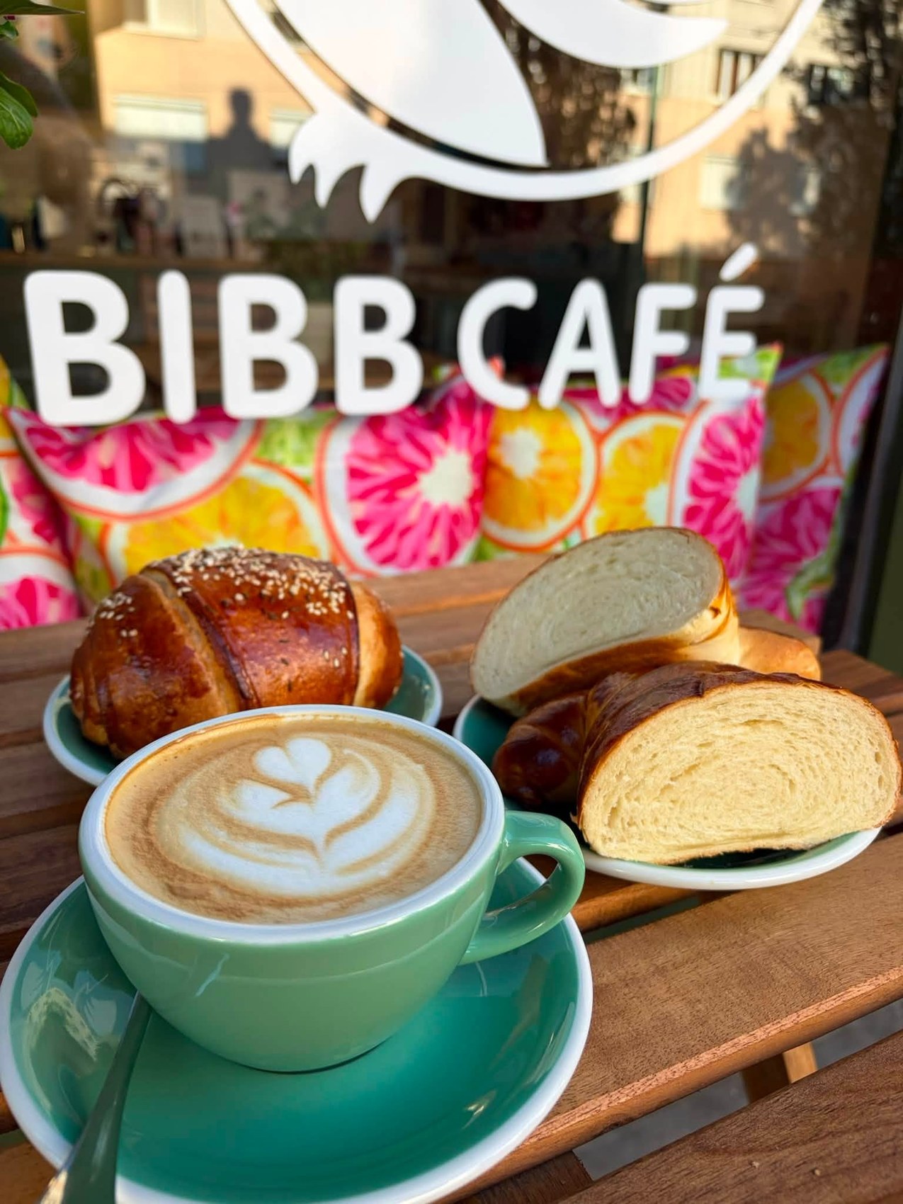 A cappuccino with heart shaped latte art next to fresh pastries on the table at BIBB Café, the shop's window lettering behind
