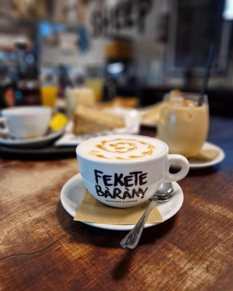 A branded Fekete Bárány cup with swirled latte art, cake and an iced drink on the table behind