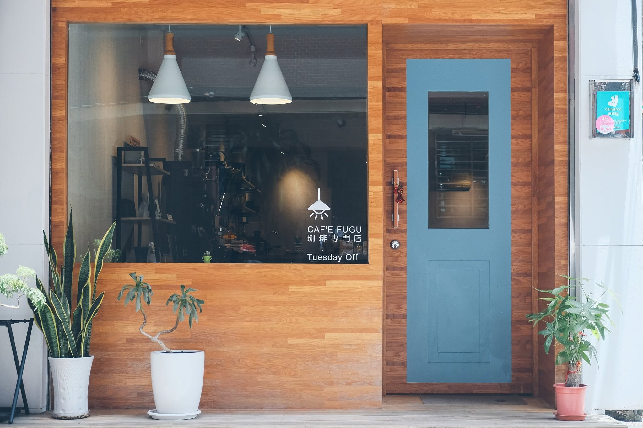 CAF'E FUGU's warm wood paneled storefront with a blue door, two pendant lamps over the window bar, and potted plants out front
