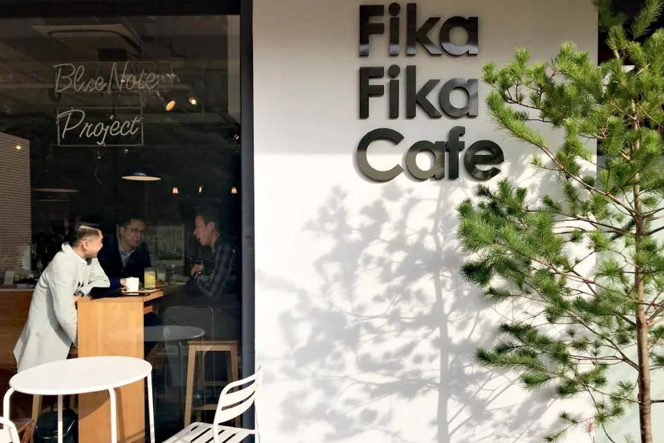 The white exterior wall of Fika Fika Cafe's Yitong Street shop with raised FIKA FIKA CAFE lettering, a young pine tree out front, and guests seated at the window bar inside