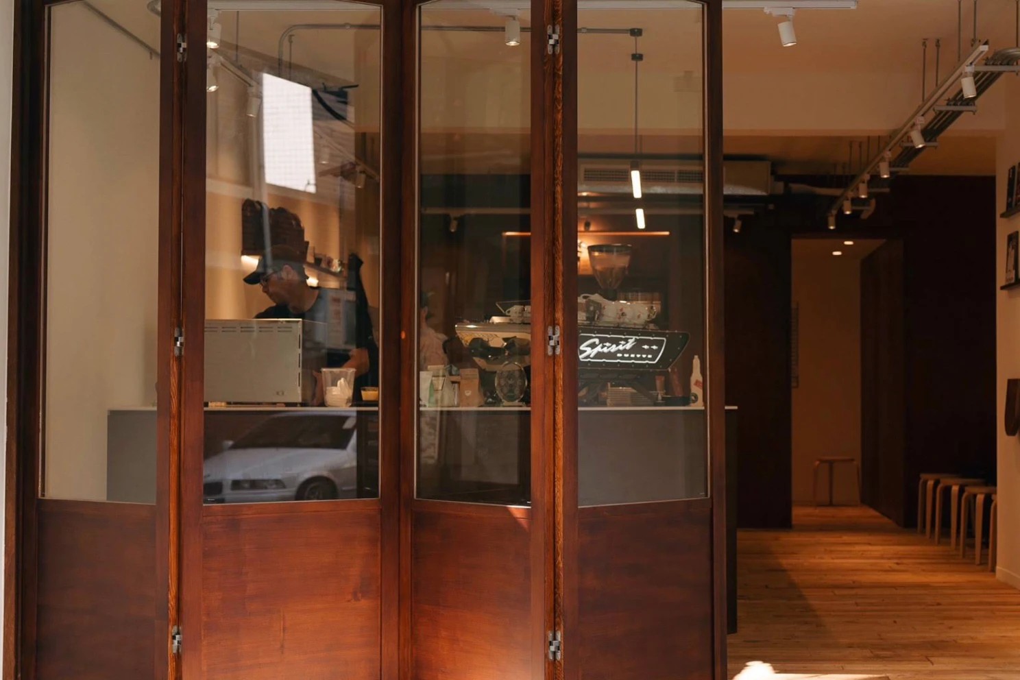 The folding wood and glass front of Kite Coffee, a barista at work inside and a Spirit espresso machine on the bar beside a row of wooden stools