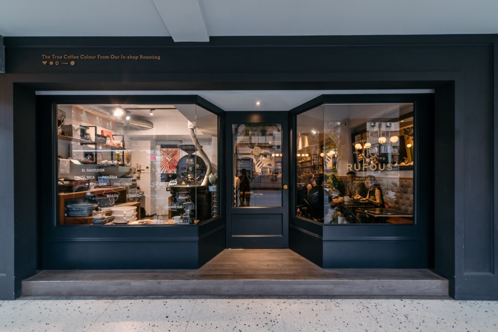 Rufous Coffee's dark navy storefront with RUFOUS lettering etched on the window glass, guests seated inside past a roaster and shelves of origin coffee sacks