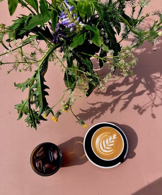 An iced coffee and a latte with heart art on a pink table beside cut flowers at Kokomo