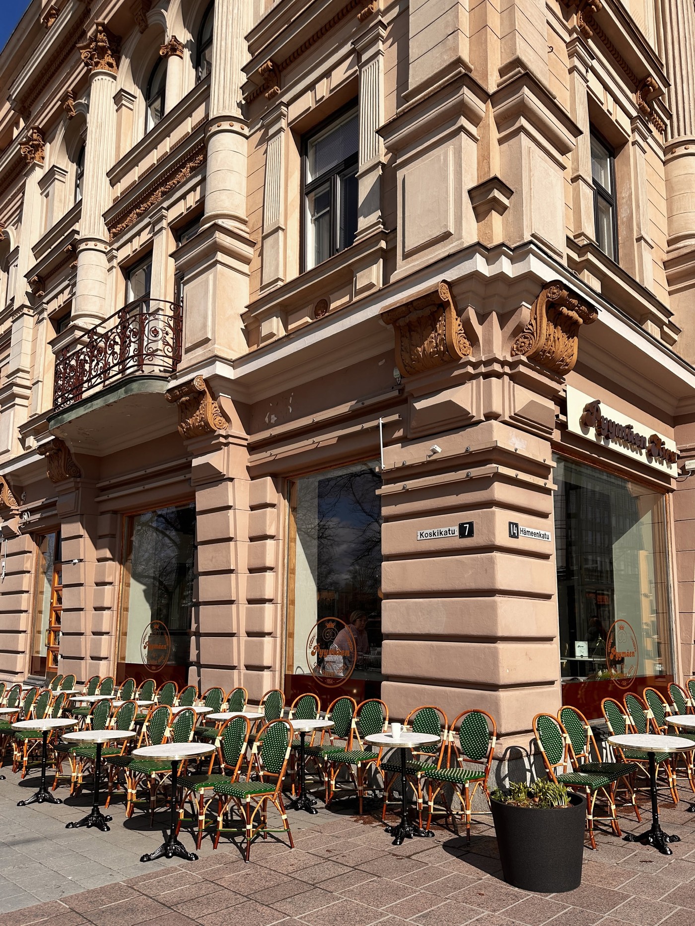 Rows of green rattan bistro chairs and marble top tables outside the ornate corner storefront of Pyymäen Aira