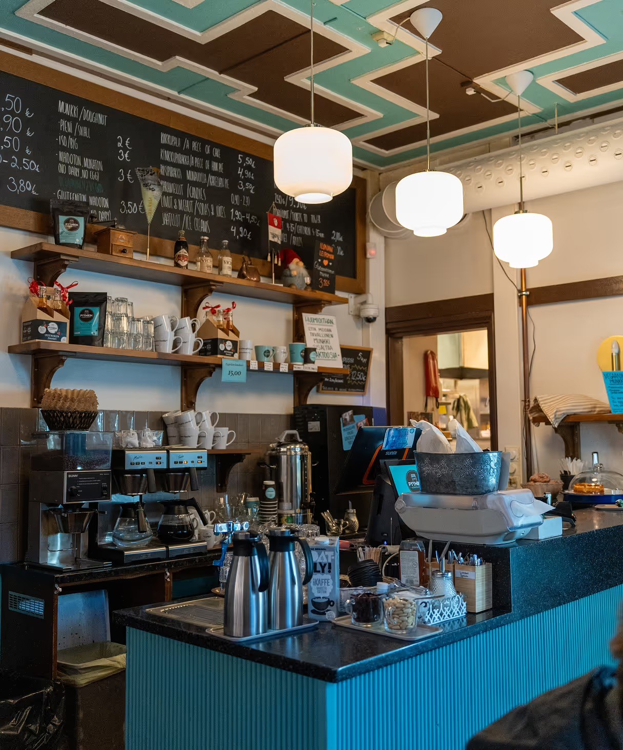 A chalkboard doughnut menu above shelves of mugs and retail coffee bags at Pyynikin Munkkikahvila's teal counter