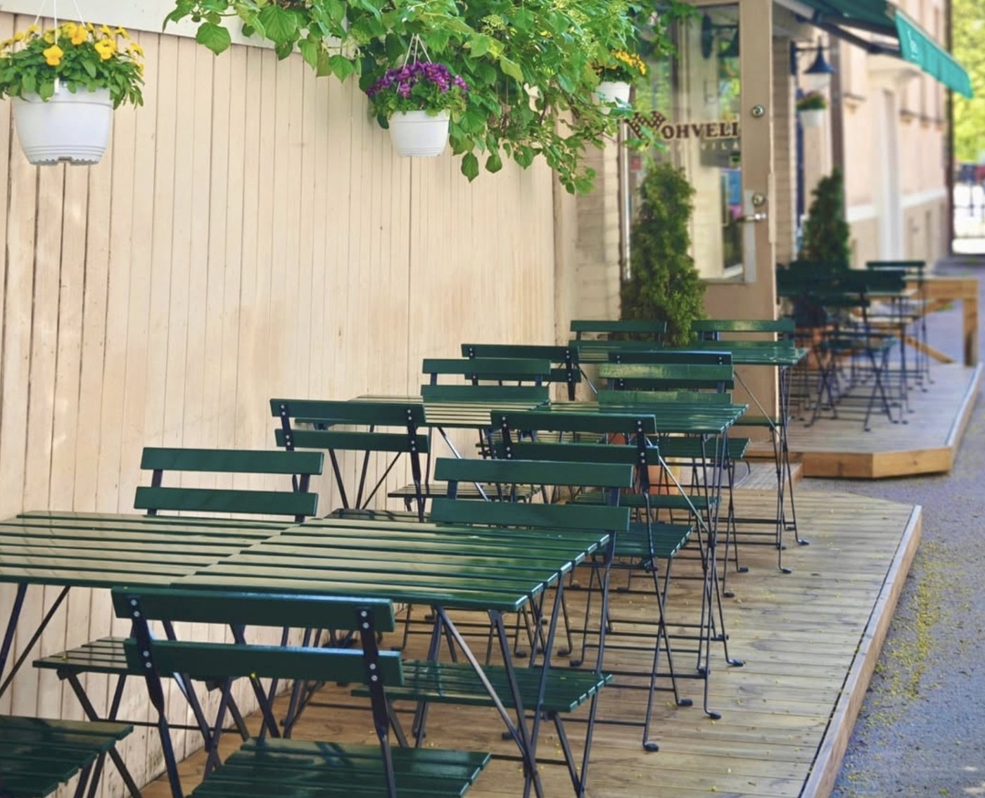 Rows of empty green bistro tables and chairs on Vohvelikahvila's wooden terrace under hanging flower baskets