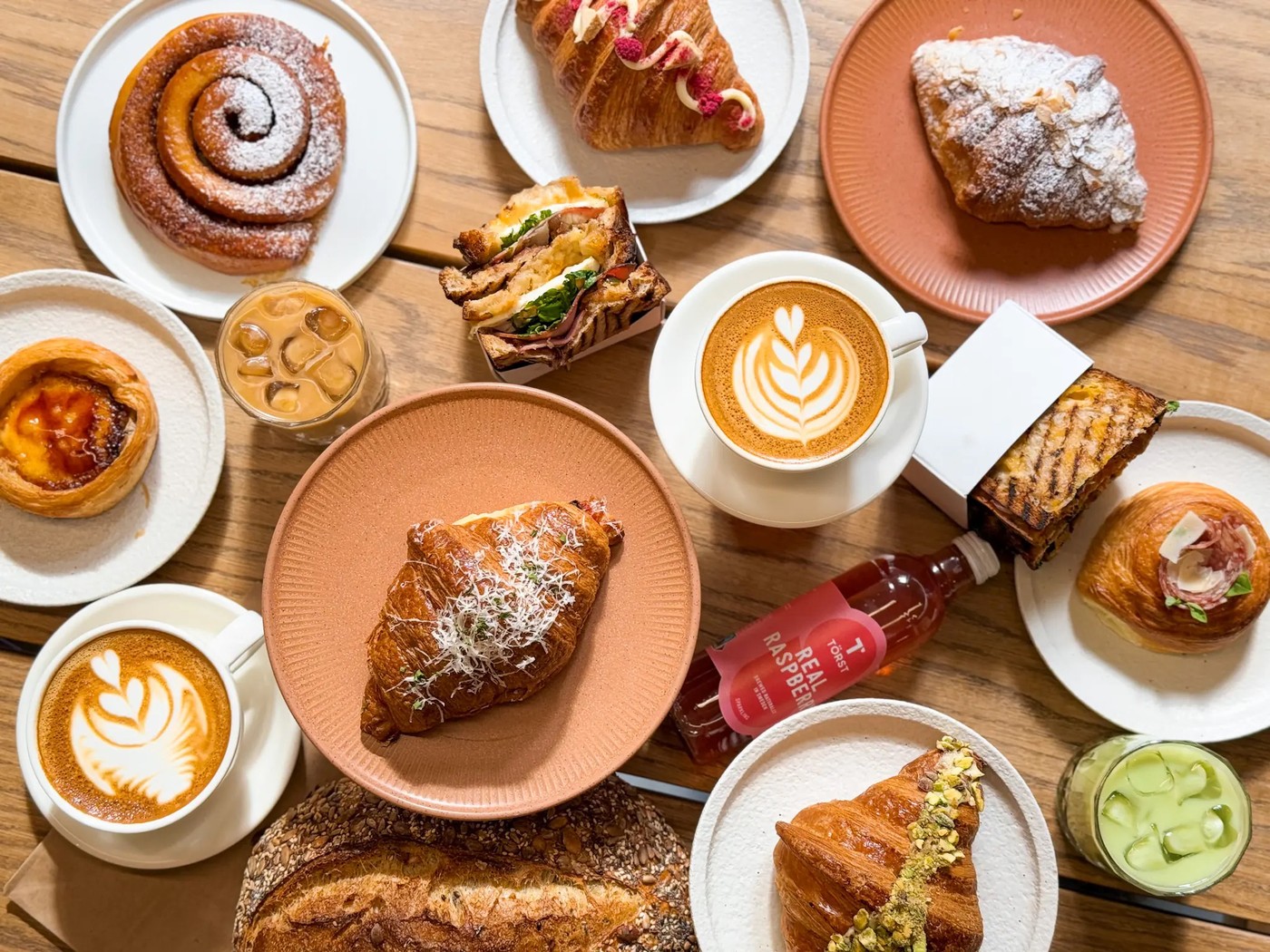 An overhead spread of pastries, croissants, a sandwich, a loaf of sourdough, and two lattes with leaf art on a wooden table