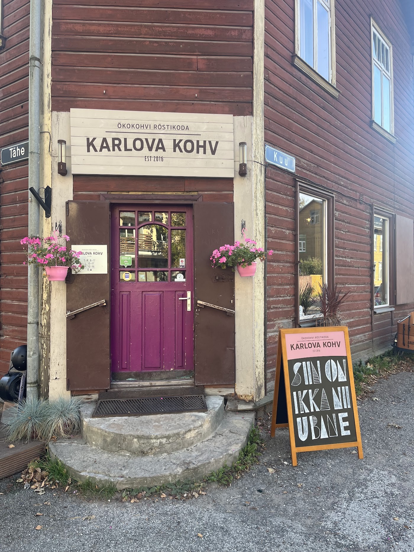Wooden house facade with a purple front door under a Karlova Kohv Est 2016 sign, pink flower baskets hanging on either side