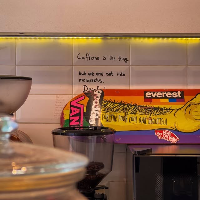 A grinder and skateboard deck against a tiled wall hand-lettered with 'Caffeine is the king but we are not into monarchs'