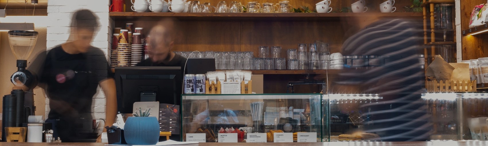 A long-exposure shot behind the counter at Kolektiva, baristas in motion against shelves of cups, glassware and bagged beans, a pastry case up front
