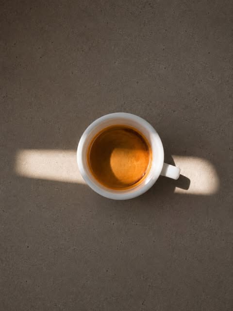 An overhead shot of a white espresso cup on a warm grey concrete surface, sunlight cutting a bright stripe across the frame