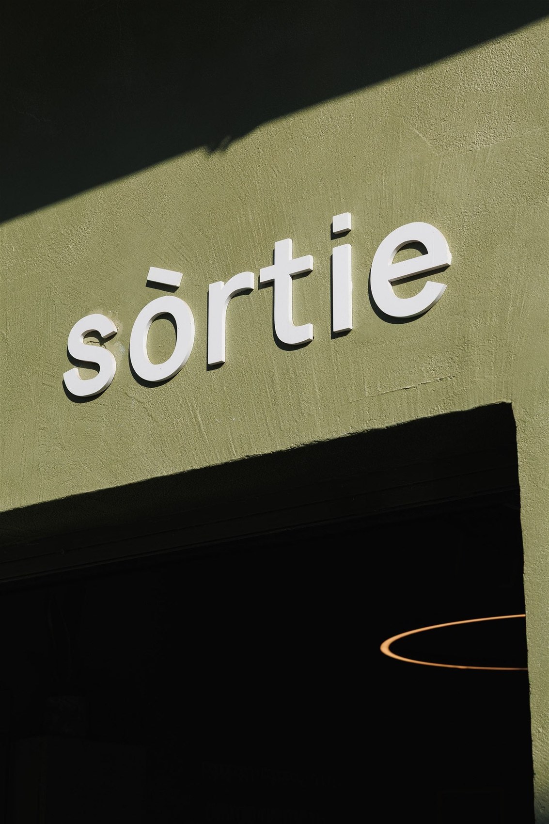 Close-up of the sōrtie storefront sign in white lettering on an olive green wall, afternoon sun cutting a sharp diagonal shadow across the facade