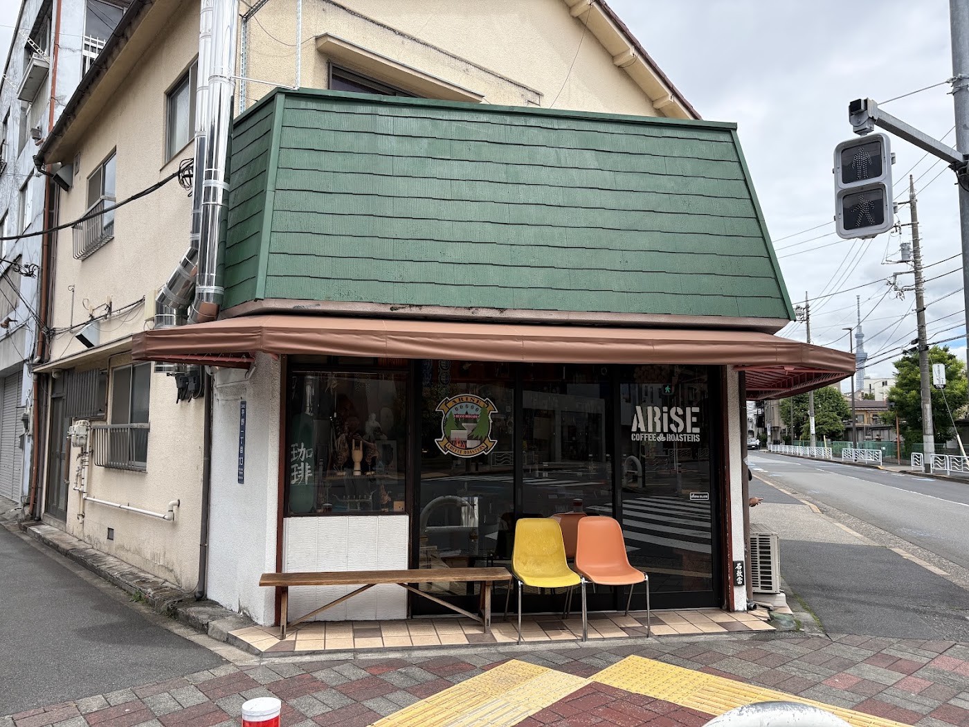 The green tiled corner storefront of Arise Coffee Roasters with colorful chairs out front and the Tokyo Skytree in the distance