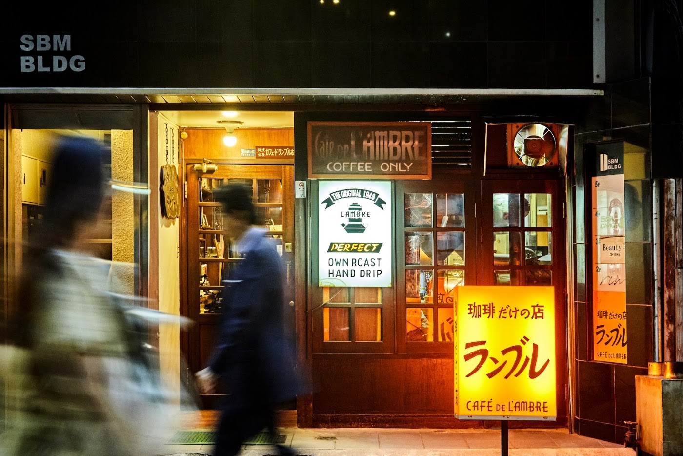 The illuminated Cafe de l'Ambre entrance at night, reading Coffee Only and The Original 1948, Own Roast Hand Drip
