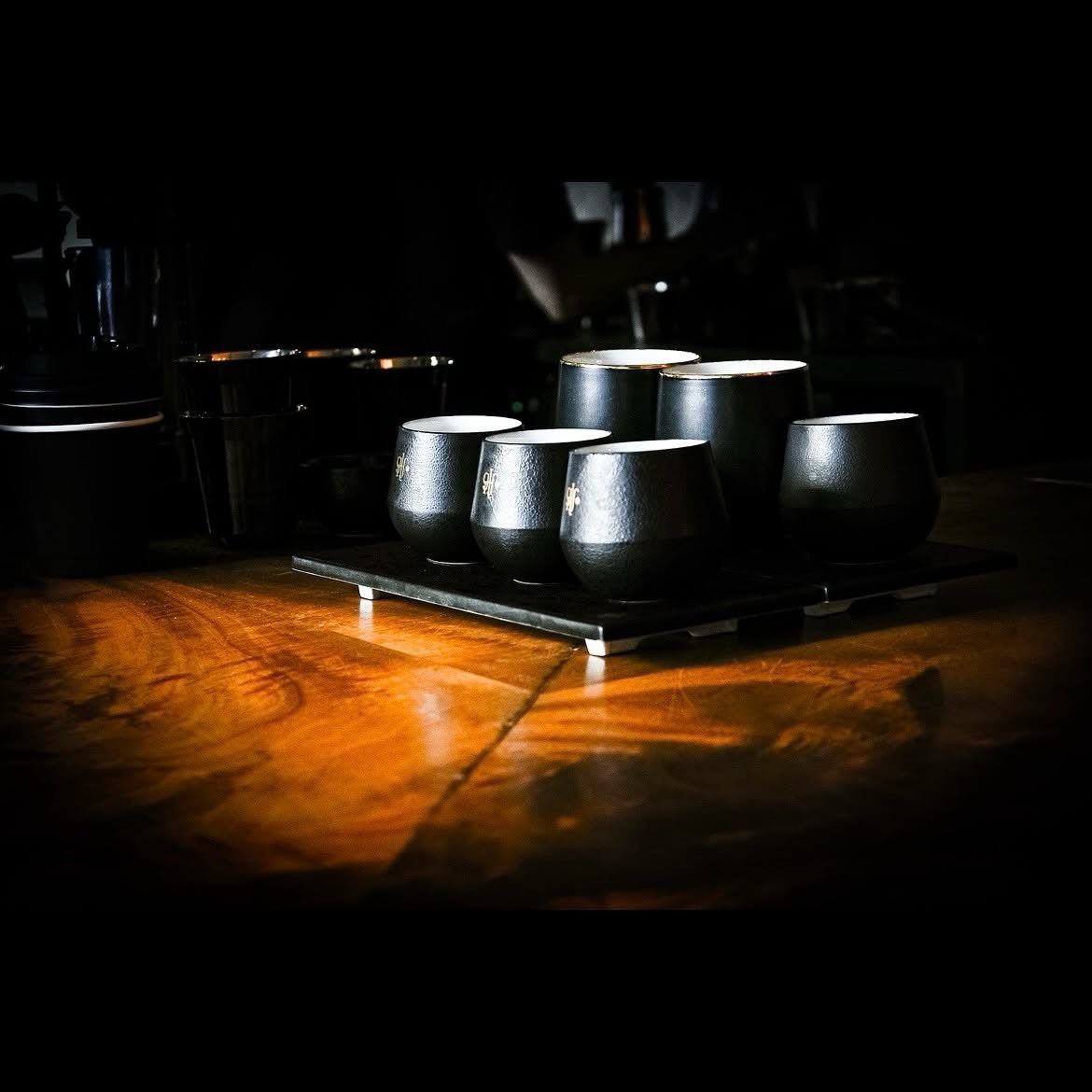 A row of black Glitch branded cupping glasses lit dramatically on the wooden counter, an espresso machine in shadow behind