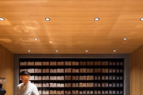 Inside Koffee Mameya, a staff member stands below the wall of labeled bean canisters beneath a warm wood ceiling