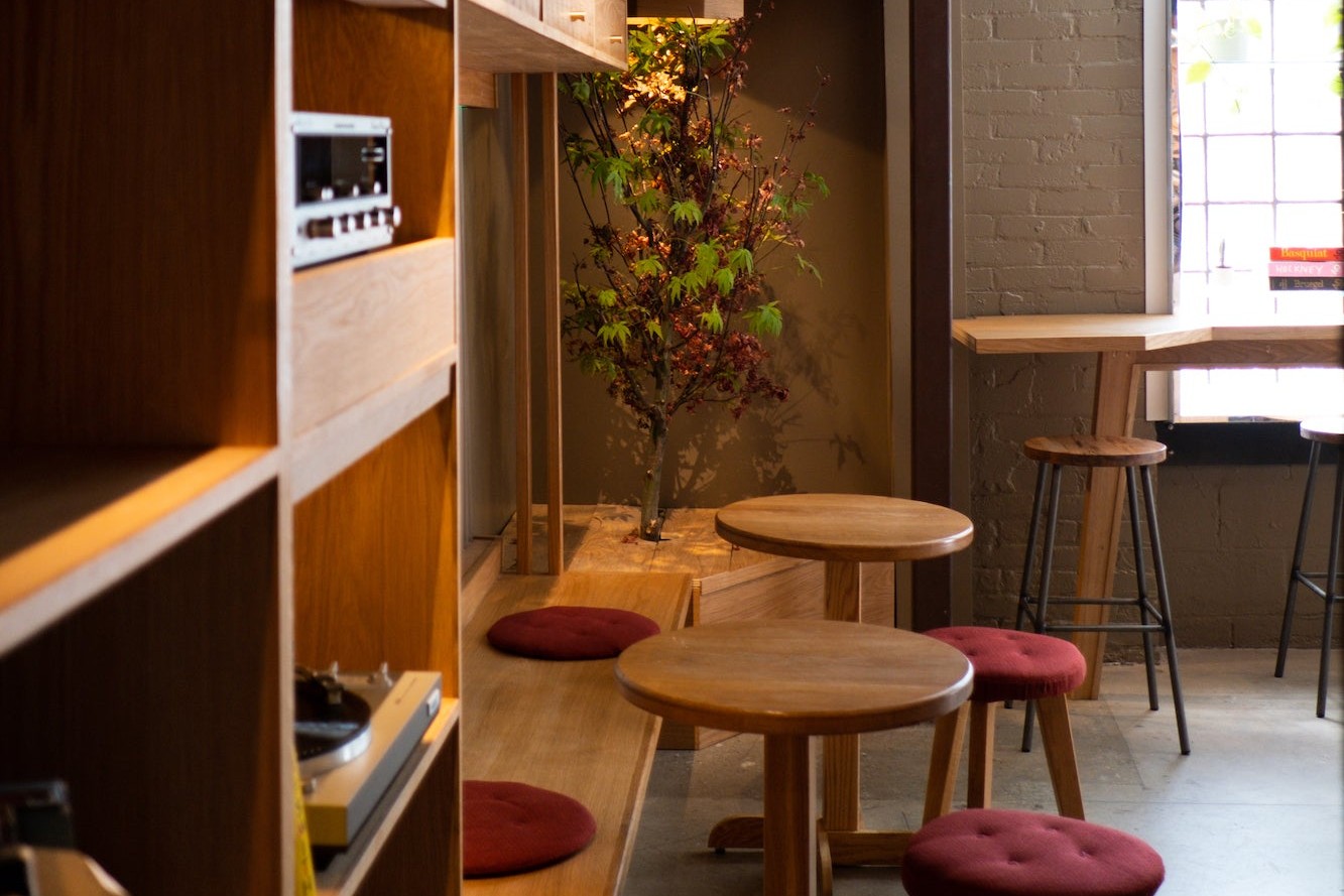 Oak shelving with a stereo receiver and turntable beside low round tables, red cushioned stools and a small tree in Boxcar Social Laneway