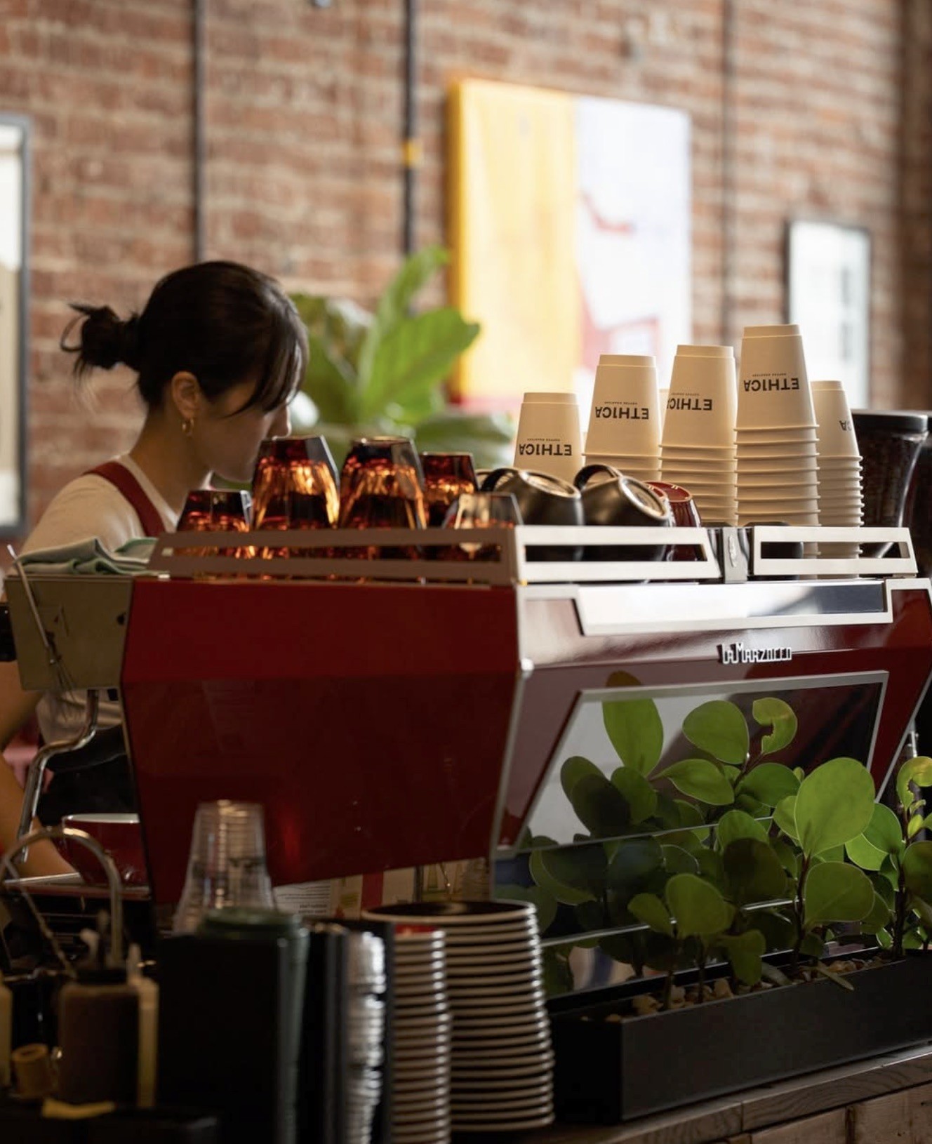 The red La Marzocco espresso machine at Ethica with stacked branded ETHICA cups, exposed brick, and potted plants behind