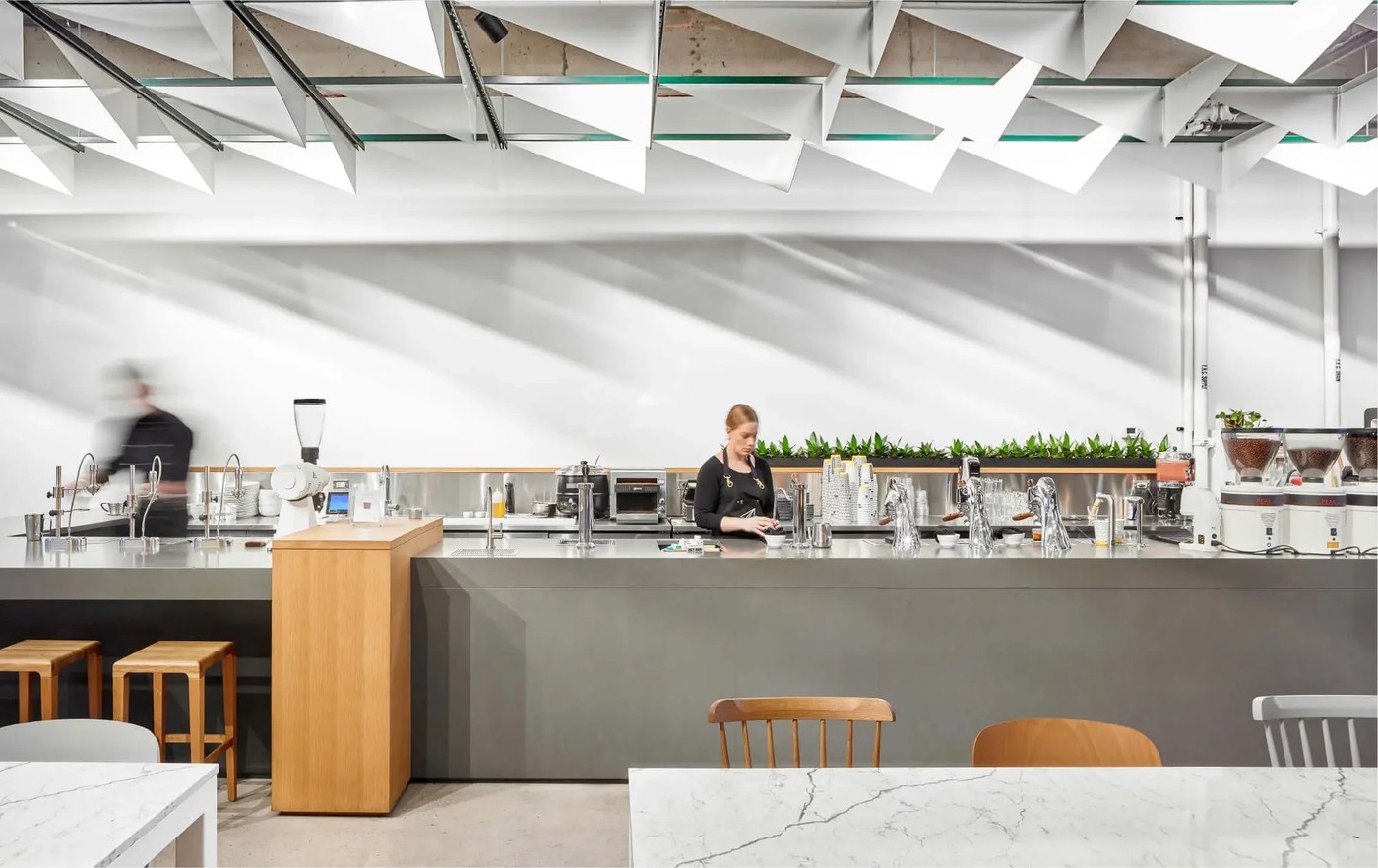 The long stainless steel bar at Pilot Coffee Roasters under an angular origami style ceiling, with rows of grinders, bean hoppers, and a barista at work