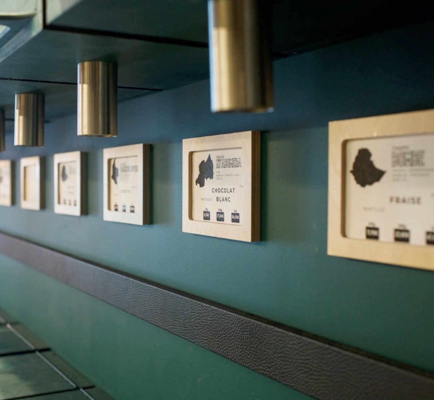 Framed origin labels for single origin beans, including a Rwanda Nyabumera chocolat blanc, mounted on a teal wall beneath brass spotlights