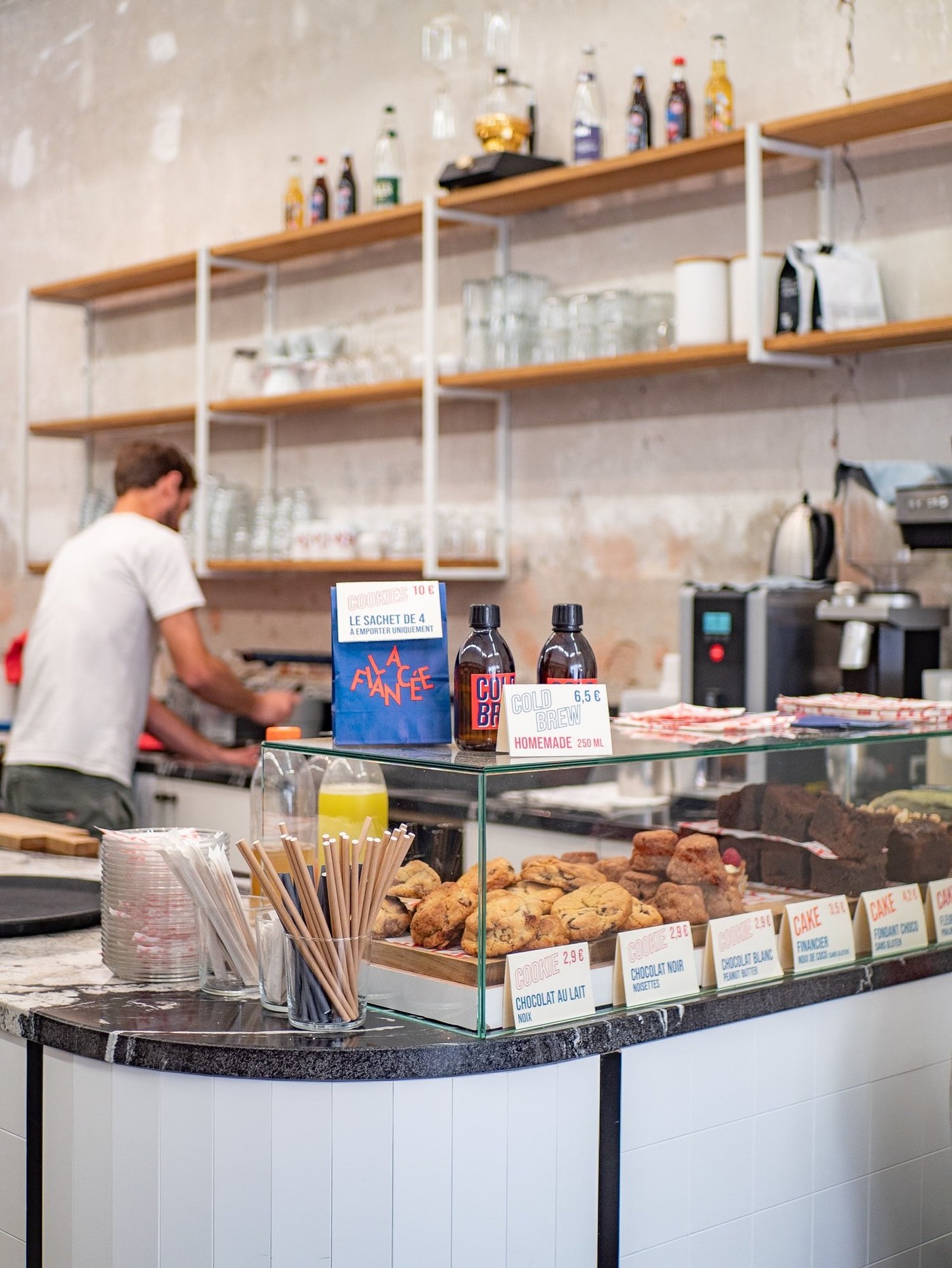 The pastry counter with cookies, brownies and bottled homemade cold brew below a shelf of glassware, a barista working with his back turned