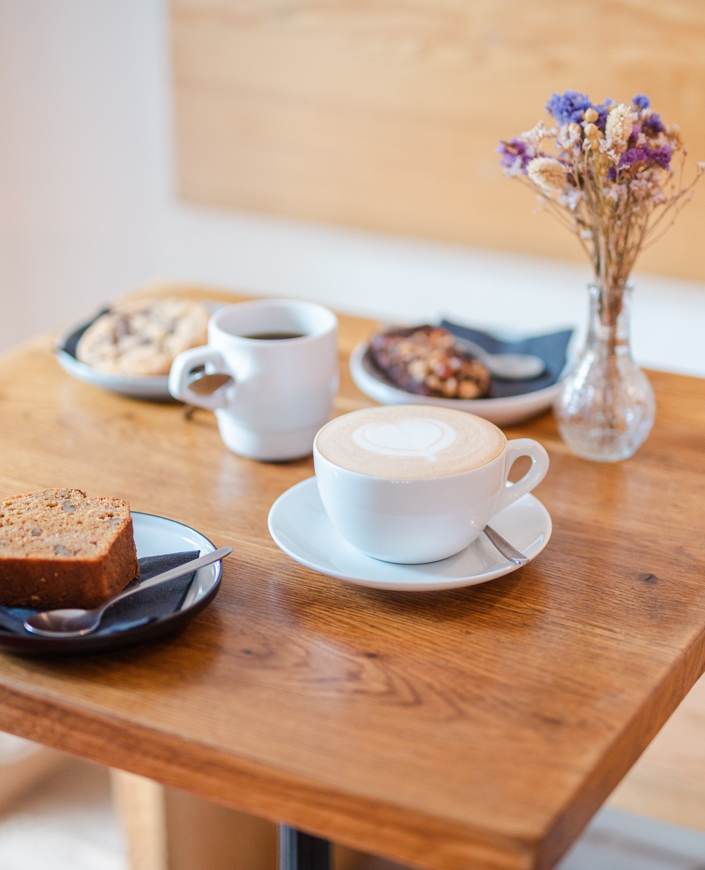 A cappuccino with heart shaped latte art on a wooden table beside a black coffee, banana bread, pastries and a small vase of dried flowers