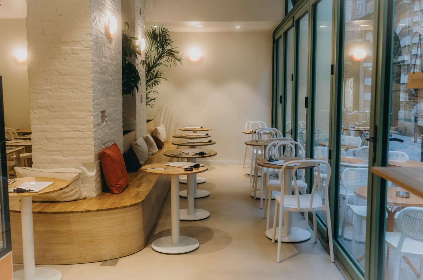 The dining room at Les Infiltres, with round wooden cafe tables, white bentwood chairs, a raw brick column and a built in bench with cushions beside floor to ceiling folding glass doors