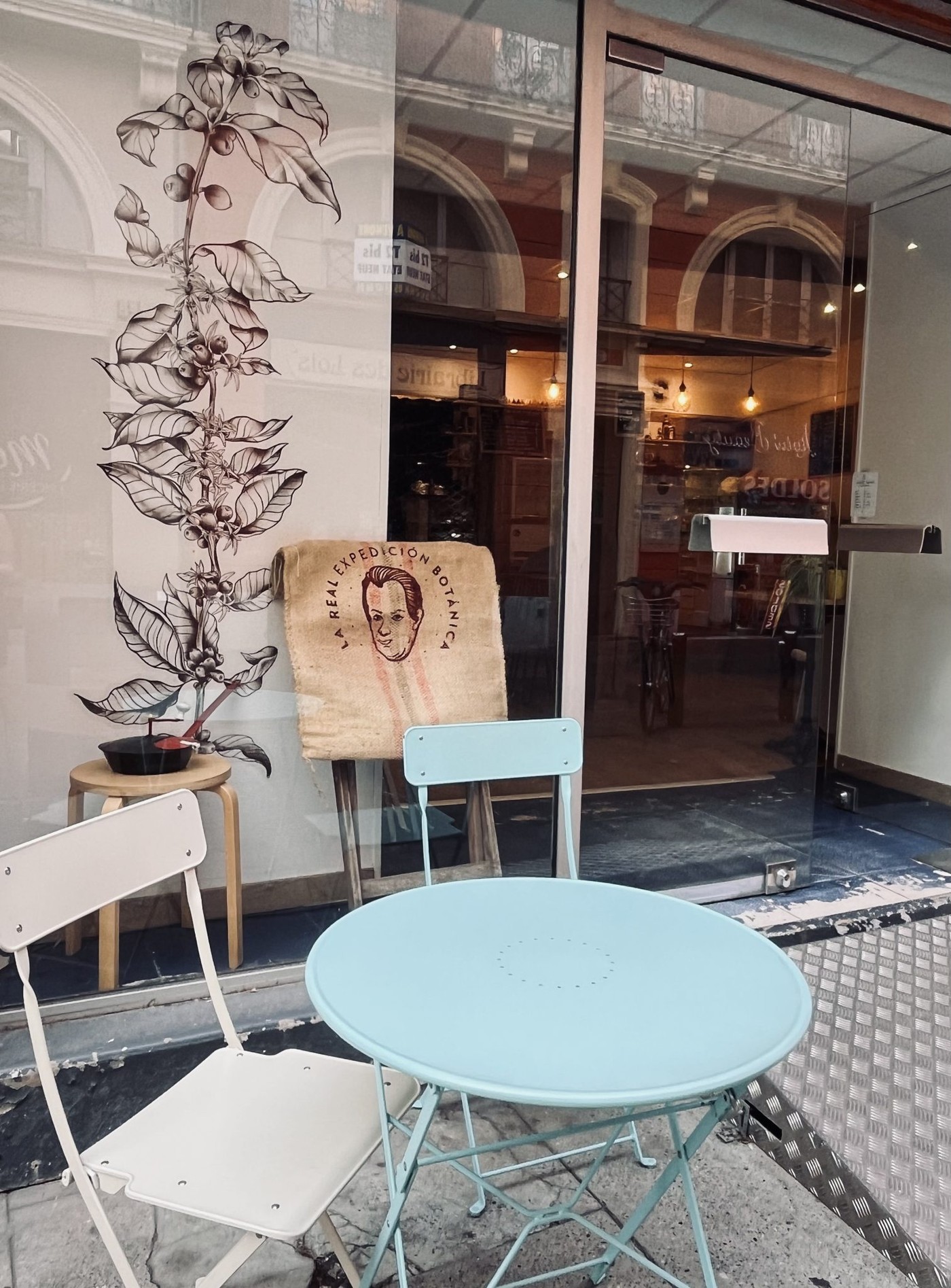 The cafe storefront window decorated with a coffee plant illustration and a burlap coffee sack on display, with a pastel outdoor table and chairs