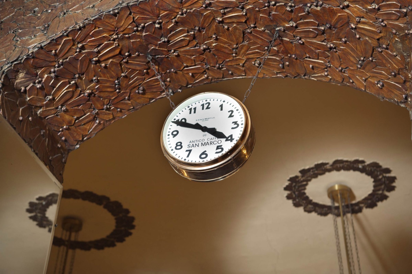 An antique brass clock reading 'Antico Caffè San Marco' hanging from an ornate carved ceiling in the Secession style