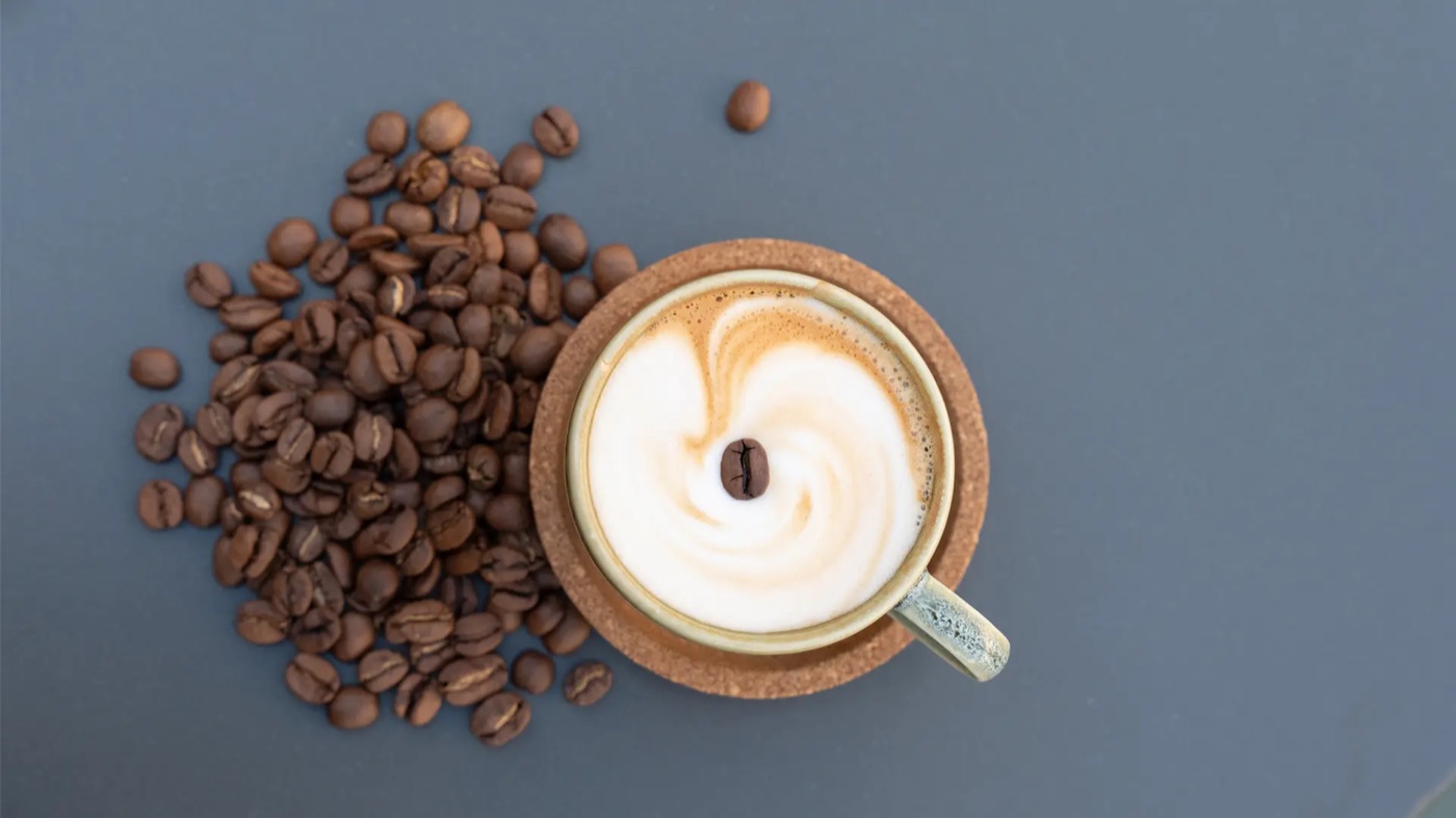 A latte with a single coffee bean garnish beside a pile of roasted coffee beans on a cork coaster
